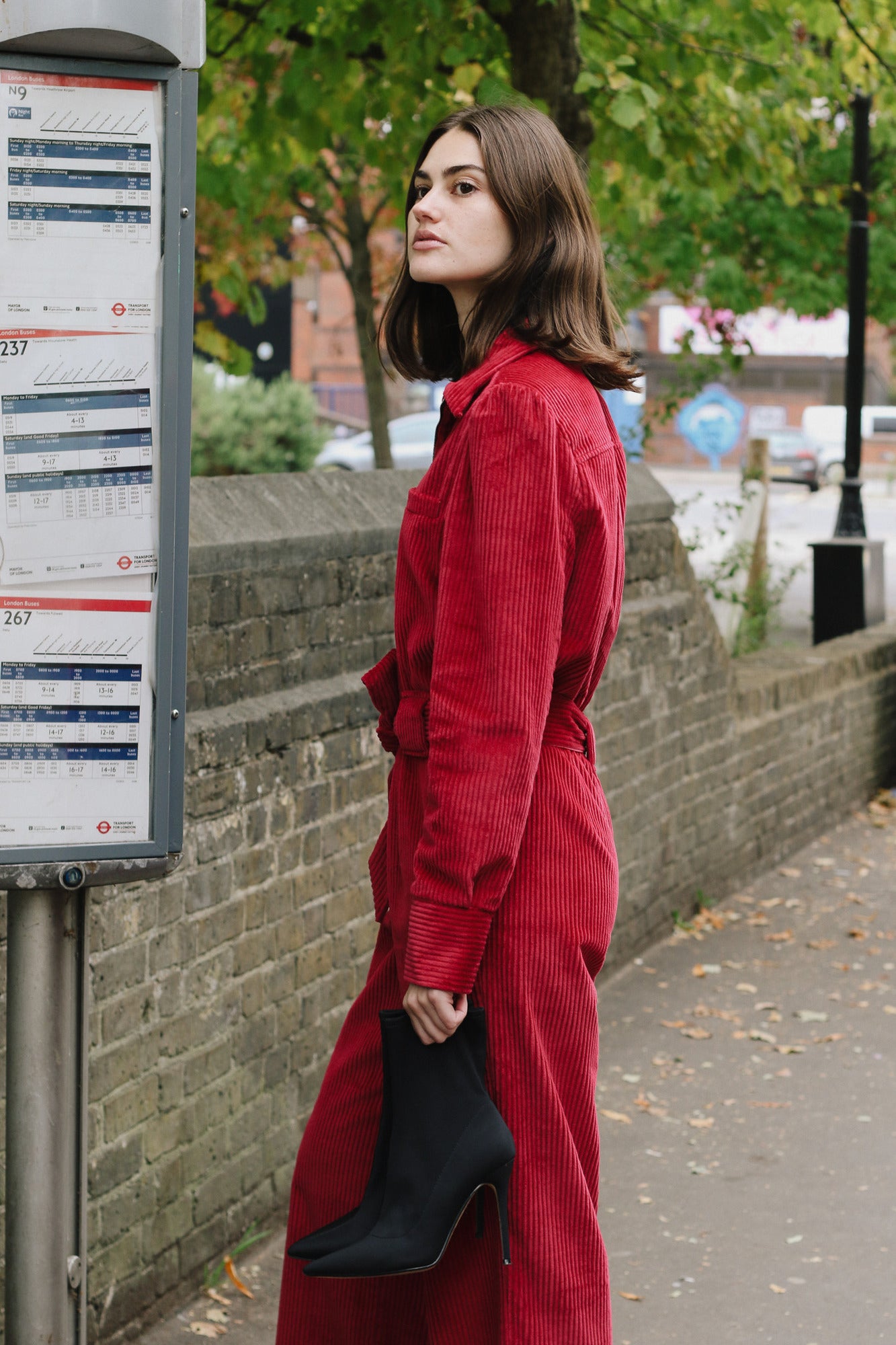 Woman in a red Winston Jumpsuit standing next to a bus stop sign with trees and buildings in the background.