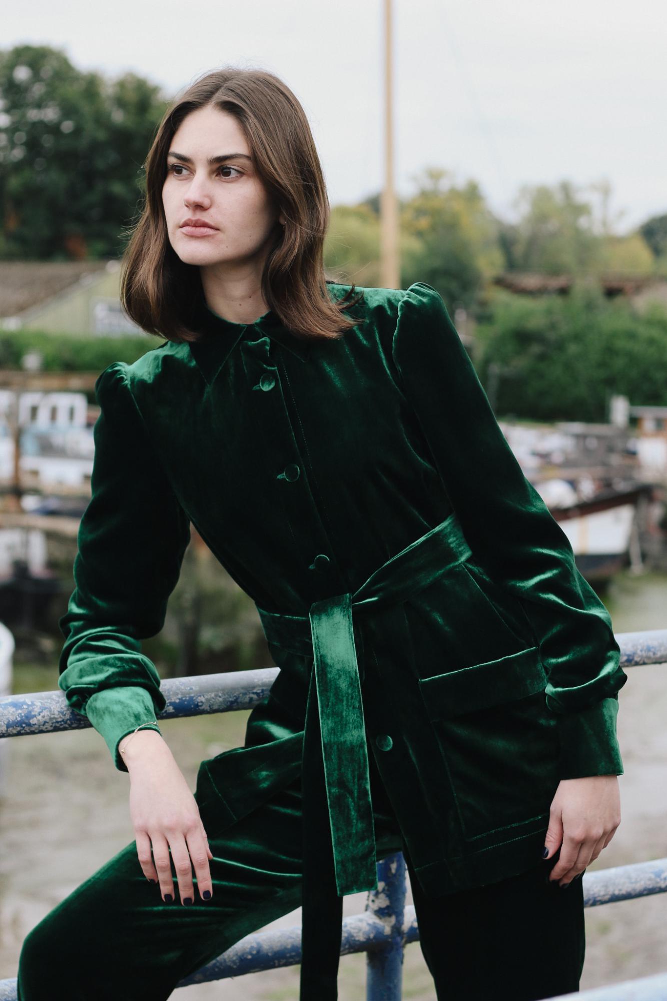 Woman wearing the velvet Gentlewomen Jacket in green leaning against gate. 