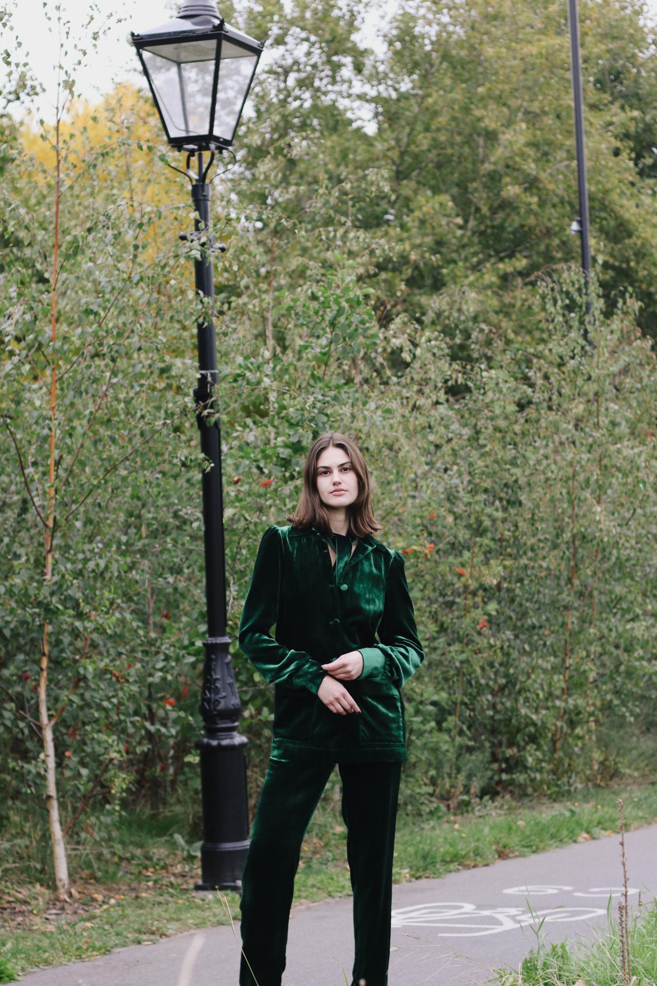 woman wearing the velvet Gentlewomen Trousers and jacket in green standing next to a lamp post with trees in the background